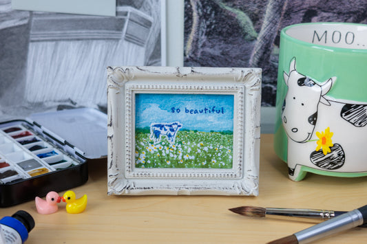 A small framed art print featuring a blue and white cow in a green field with the text 'so beautiful' sits on a wooden desk surrounded by painting supplies, including paintbrushes, a watercolor palette, and a green ceramic mug.