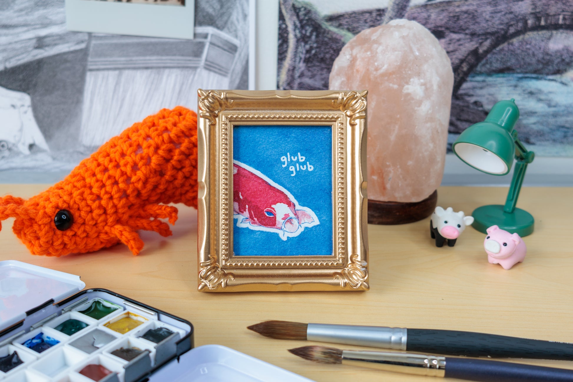 A framed art print featuring a red koi fish on a blue background with the words 'glub glub' in white, styled on a desk with a gold frame, a crocheted orange fish, paintbrushes, a watercolor palette, and a pink salt lamp.
