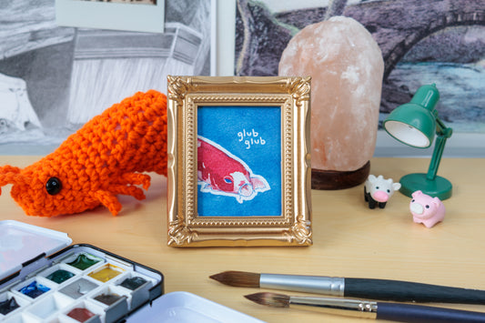 A framed art print featuring a red koi fish on a blue background with the words 'glub glub' in white, styled on a desk with a gold frame, a crocheted orange fish, paintbrushes, a watercolor palette, and a pink salt lamp.