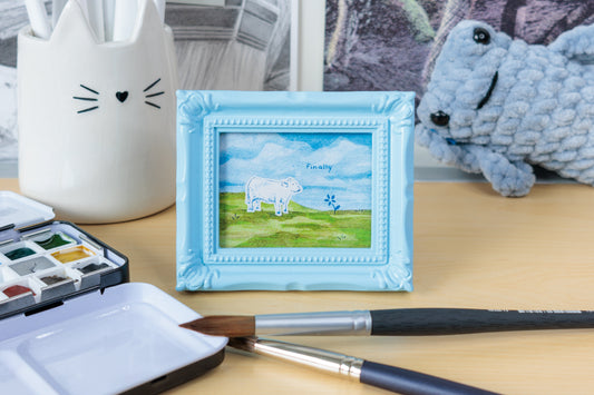 A small framed watercolor painting of a white sheep in a green field under a blue sky, displayed on a wooden desk with paintbrushes, a watercolor palette, and a white cat-shaped paint holder in the background.