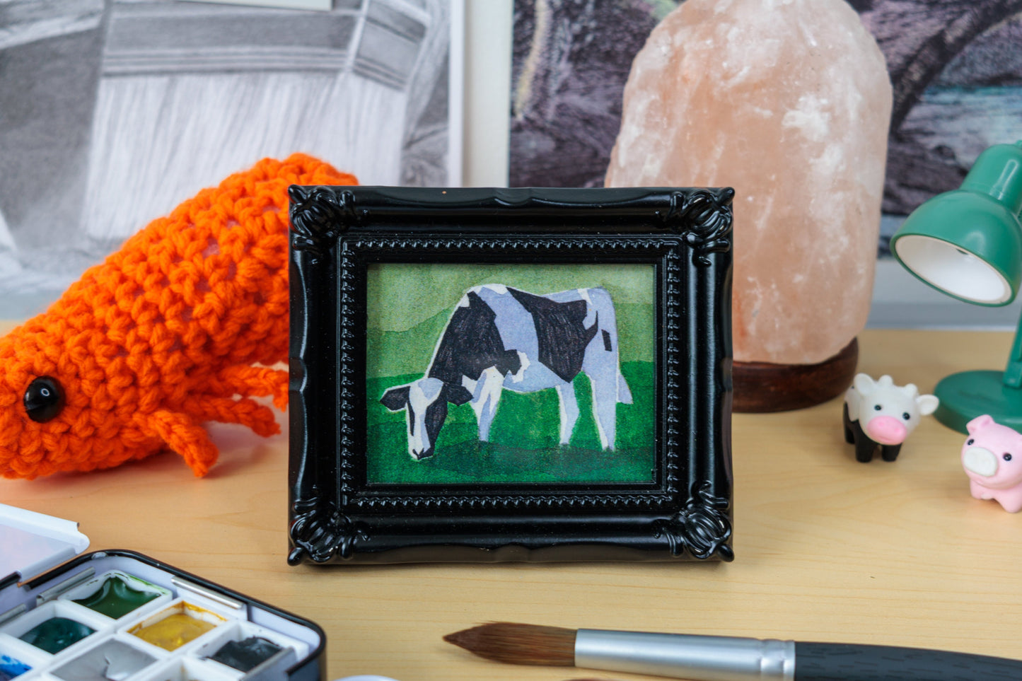 A framed miniature painting of a black and white cow on a green background, displayed on a wooden desk surrounded by art supplies, a crocheted orange animal, a pink salt lamp, and a green desk lamp.
