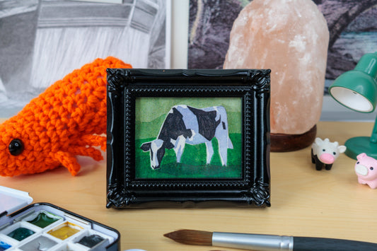 A framed miniature painting of a black and white cow on a green background, displayed on a wooden desk surrounded by art supplies, a crocheted orange animal, a pink salt lamp, and a green desk lamp.