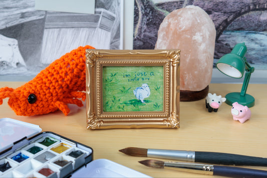 A styled tabletop scene featuring a small gold-framed painting of a white chick on green grass with the text 'I'm just a little guy.' Surrounding the frame are an orange crocheted fish, a pink salt lamp, a green desk lamp, small pig figurines, paintbrushes, a watercolor palette, and a sketchbook in the background.