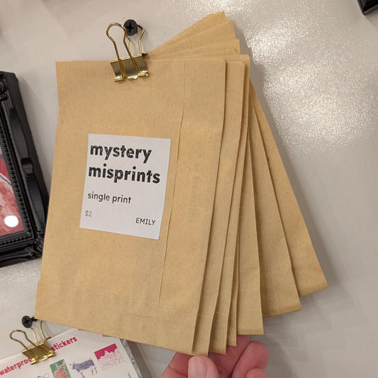 Hand holding a stack of brown paper bags with 'mystery misprints' label on a surface with stationery items.